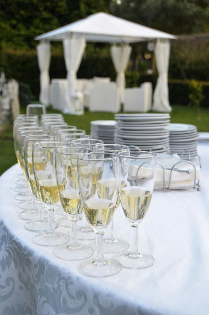 A table set with champagne flutes filled with sparkling wine, in front of a white tent and serving plates in an outdoor setting.