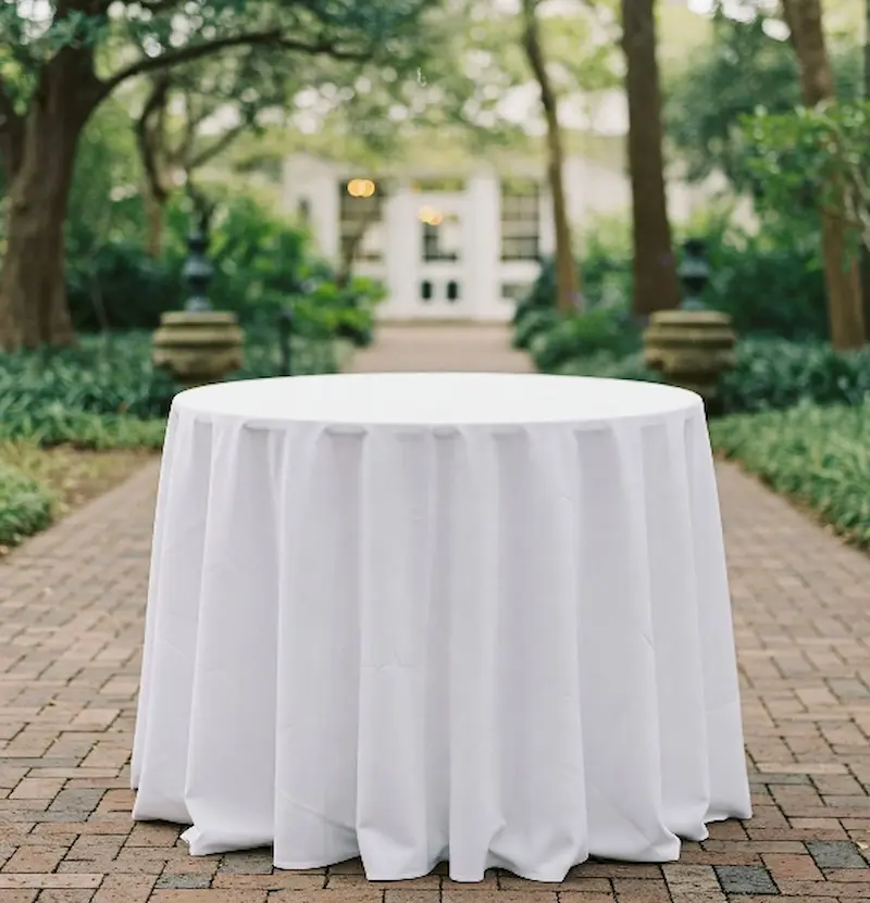 A round table covered with a white tablecloth, set in a garden pathway with lush greenery and a building in the background.
